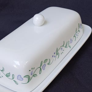 May include: A white ceramic butter dish with a rectangular base and a matching lid featuring a round knob handle. The lid is decorated with a repeating pattern of green leafy vines, small purple dots, and light purple heart-shaped accents.