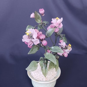 May include: A decorative bonsai tree in a light green ceramic pot. The tree features pink flowers with yellow centers, green leaves, and small pink buds. The pot is filled with white pebbles, creating a serene and elegant aesthetic.