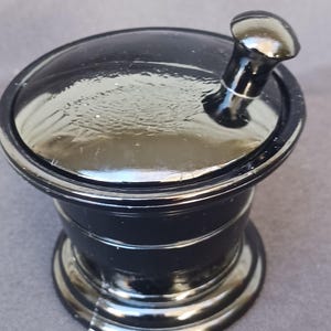 May include: A black mortar and pestle set. The mortar is a cylindrical shape with a wide base and a flared rim. The pestle has a rounded head and a short, thick handle. The set is made of a glossy, dark material.