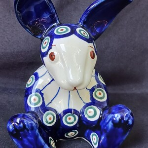 May include: A ceramic rabbit figurine featuring a glossy white base with deep blue patches and circular patterns in green, white, and orange. The rabbit is in a seated position with long ears, a smooth finish, and distinct painted eyes.