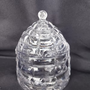 May include: A clear, glass jar with a domed lid. The jar and lid are decorated with a diamond pattern. The lid has a small, round knob on top. The jar is likely used for storing small items or as a decorative piece.