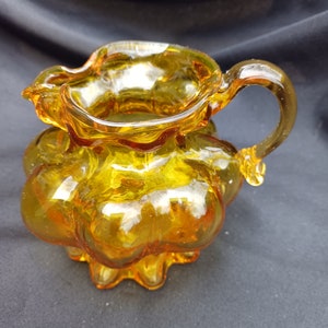 May include: A small amber glass pitcher with a ruffled rim and a bulbous body. The pitcher has a single handle and a wavy base.