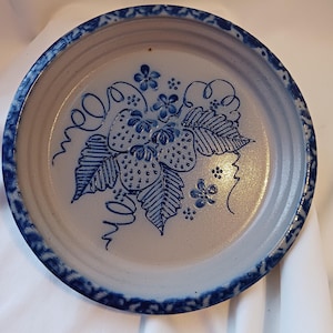 May include: A round, ceramic plate with a light beige center and a dark blue rim. The plate features a hand-painted design of strawberries and leaves in blue. The rim has a decorative blue pattern.