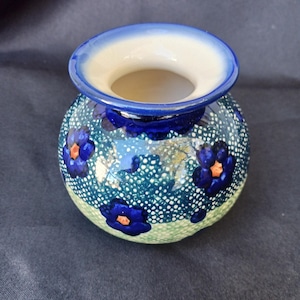 May include: A small, round ceramic vase with a blue and white floral pattern. The vase has a wide opening with a white interior and a blue rim. The exterior features blue flowers with orange centers on a teal background.