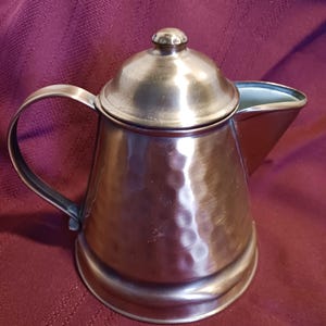 May include: A vintage silver-toned metal coffee pot with a hammered texture. The pot features a curved handle, a spout, and a lid with a small knob. The pot is likely made of stainless steel or a similar metal. The background is a deep red fabric.