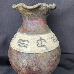 May include: A handcrafted ceramic vase with a ruffled rim. The vase features a textured, iridescent finish in shades of burgundy, brown, and green. A cream-colored band with etched symbols encircles the vase, adding a touch of artistry.