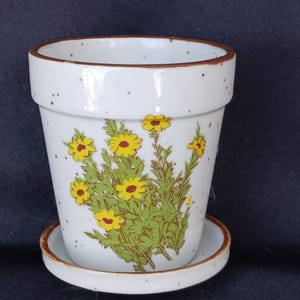 May include: White ceramic flower pot with a matching saucer. The pot features a floral design with yellow flowers and green leaves. The rim and base have a brown trim. The pot is approximately 5 inches tall and 5 inches in diameter.