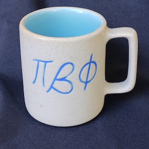 May include: A speckled ceramic mug with a light blue interior and a large handle. The exterior is a speckled off-white color, and the mug features blue Greek letters. The mug is set against a dark blue background.