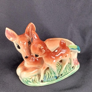 May include: A ceramic planter shaped like a mother and baby deer. The deer are brown with white spots and have black eyes. The planter is decorated with green and blue leaves.