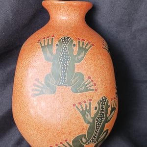 May include: A rounded ceramic vase with a speckled orange finish, featuring two green frog illustrations with white dotted backs and red-tipped toes. The vase has a narrow neck and a smooth, bulbous body, showcasing a detailed, hand-painted design.