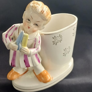 May include: A vintage porcelain figurine of a young child holding a comb and brush, next to a white container with floral accents. The child wears a white outfit with purple stripes and orange shoes. The container has gold trim.