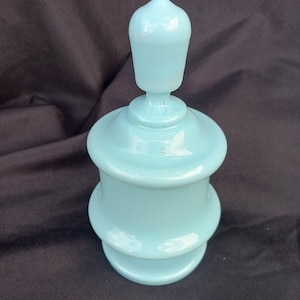 May include: A light blue glass bottle with a rounded top and a decorative design. The bottle has a smooth, glossy finish.