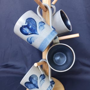 May include: A set of three ceramic mugs with a speckled light gray exterior and a dark interior, each featuring a hand-painted blue heart design. The mugs are displayed on a light-colored wooden stand with multiple pegs for hanging.