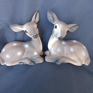 May include: Two ceramic deer figurines. The deer are light brown with white spots and red eyes. They are in a resting position, with their legs tucked beneath them. The background is a dark blue fabric.