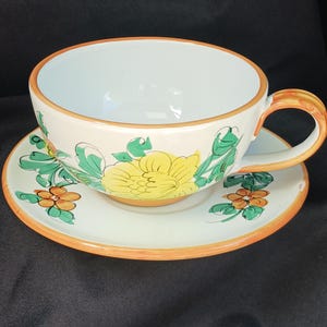 Könnte beinhalten: Ein weißes Keramik-Tee-Tassen- und Untertassen-Set. Die Tasse und die Untertasse sind mit gelben und orangefarbenen Blumenmustern und grünen Blättern verziert. Die Ränder und der Griff sind orange. Die Teetasse steht auf einer passenden Untertasse.