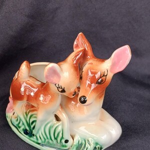 May include: A ceramic planter shaped like a mother and baby deer. The mother deer is laying down with her head turned to the side and her eyes closed. The baby deer is standing on its hind legs and looking at the mother deer. The planter is painted in shades of brown, white, and green.