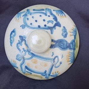 May include: A round ceramic lid with a cream-colored base featuring hand-painted blue cow illustrations, blue polka dots, and small yellow floral accents. The lid has a central rounded knob handle and a smooth, glossy finish throughout the surface.