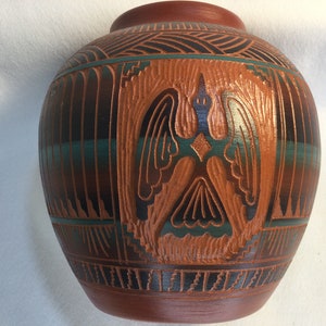 May include: A brown ceramic pot with a carved design of a bird with outstretched wings. The pot is decorated with black and turquoise lines.