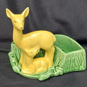 May include: A ceramic planter featuring a yellow deer and fawn figurine. The deer stands above the fawn, which is lying down. The planter has a green, rectangular container and a base with a leaf design.