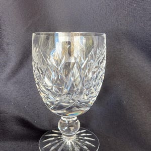 May include: A clear, cut crystal wine glass with a round base and a stem. The glass features a detailed diamond and leaf pattern, reflecting light. The base has a starburst design.