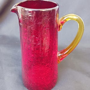 May include: A tall, ruby red glass pitcher with a textured surface and a yellow handle. The pitcher has a spout for easy pouring. The handle is a contrasting yellow color. The pitcher is likely used for serving beverages.