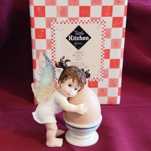 May include: A figurine of a fairy embracing a large egg in an egg cup. The fairy has brown hair, a white dress, and iridescent wings. The egg is light brown, and the egg cup is white with a blue stripe. A box with a red and white checkered pattern is in the background.