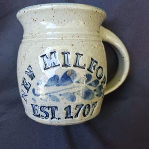 May include: A beige ceramic mug with a speckled finish and a curved handle. The front features embossed blue text reading NEW MILFORD and EST. 1707, with a blue leaf design centered between the two lines of text on the rounded body of the mug.