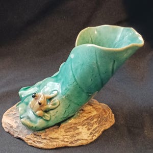 May include: A ceramic frog figurine perched on a green leaf-shaped vase. The vase is teal with a textured surface, and the frog is tan with black eyes. The base is a brown, textured, organic shape.