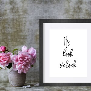 May include: A black framed print with the text "It's book o'clock" in black script font. A vase with pink roses sits in front of the print.