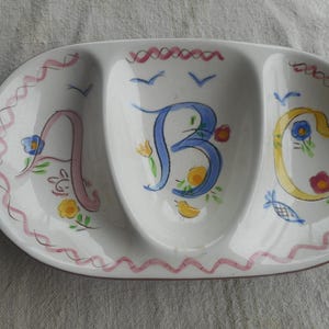 May include: An oval white ceramic divided serving dish with three sections. The dish features hand-painted letters A, B, and C in pink, blue, and yellow, accented with small floral motifs, birds, and a wavy pink border along the outer rim.