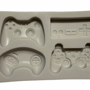 May include: A silicone mold for making cake decorations in the shape of four different video game controllers. The mold is gray and has four cavities: a classic NES controller, a PlayStation controller, an Xbox controller, and a Sega Genesis controller.