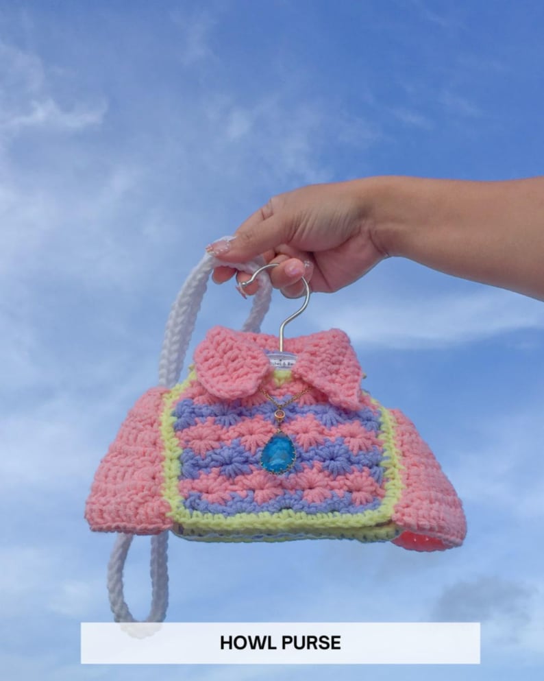 Puede incluir: Un bolso de ganchillo rosa y azul con forma de chaqueta, con una correa blanca y un colgante azul. El bolso tiene un ribete amarillo y est&aacute; sujeto por una percha plateada. El texto "HOWL PURSE" est&aacute; en la parte inferior.