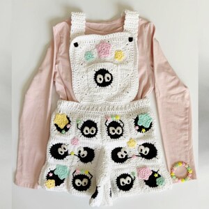 Puede incluir: Mono blanco de ganchillo con dise&ntilde;os negros de Soot Sprite, detalles de estrellas y flores de colores, y una pulsera a juego. El mono se combina con una camiseta de manga larga rosa claro. El texto en la imagen dice "SOOT SPRITES OVERALLS."