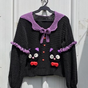Puede incluir: Su&eacute;ter negro de crochet con cuello y detalles morados. El su&eacute;ter presenta dos caras de gato con pajaritas rojas y botones en forma de coraz&oacute;n. El texto "JIJI PULLOVER" est&aacute; en la parte inferior.