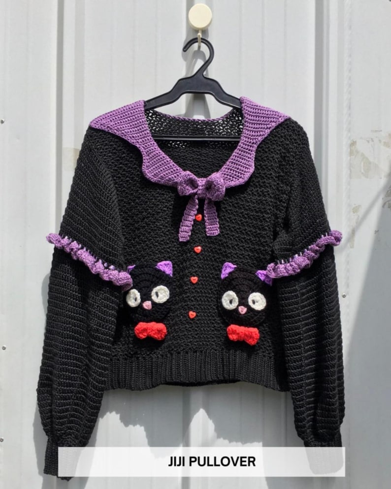 Puede incluir: Su&eacute;ter negro de crochet con cuello y detalles morados. El su&eacute;ter presenta dos caras de gato con pajaritas rojas y botones en forma de coraz&oacute;n. El texto "JIJI PULLOVER" est&aacute; en la parte inferior.