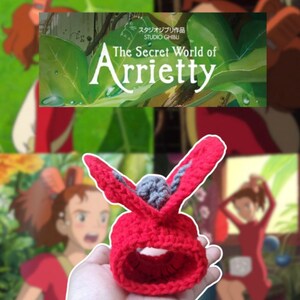 Puede incluir: Un coletero o pinza para el pelo rojo de ganchillo con detalles grises, con forma de orejas de conejo, se sostiene en una mano. El texto "ARRIETTY HAIR CLIP/CROCHET SRUNCHIE" es visible. El fondo presenta im&aacute;genes de "Arrietty y el mundo de los diminutos".