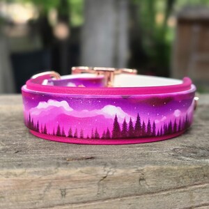 May include: A vibrant pink dog collar with a painted landscape design. The collar features a forest silhouette against a purple and pink sky, with a white interior and rose gold hardware. The collar is set on a wooden surface.