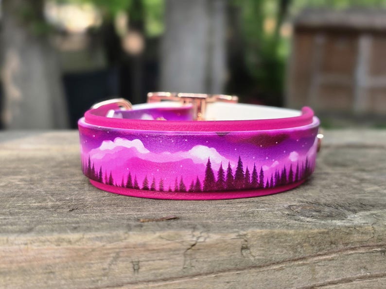 May include: A vibrant pink dog collar with a painted landscape design. The collar features a forest silhouette against a purple and pink sky, with a white interior and rose gold hardware. The collar is set on a wooden surface.
