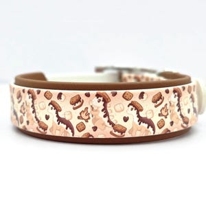 May include: A dog collar with a repeating pattern of a cartoon dinosaur with a s'more on its head, marshmallows, and graham crackers. The collar has a brown exterior, a white interior, and a white plastic buckle.
