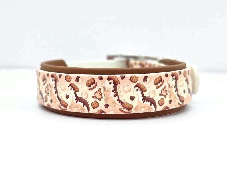 May include: A dog collar with a repeating pattern of a cartoon dinosaur with a s'more on its head, marshmallows, and graham crackers. The collar has a brown exterior, a white interior, and a white plastic buckle.