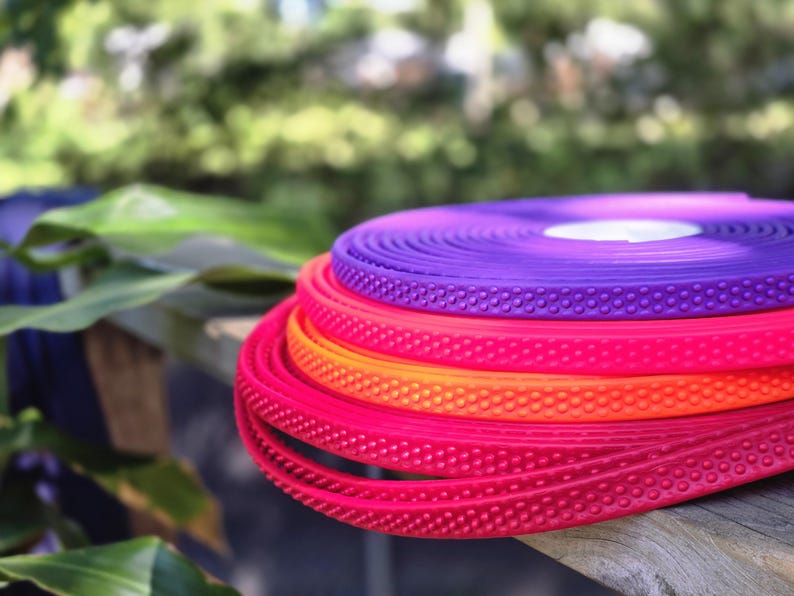 May include: A stack of colorful, coiled rubber strips in shades of red, orange, and purple. Each strip has a textured, dotted surface. The strips are resting on a wooden surface, with a blurred green background.