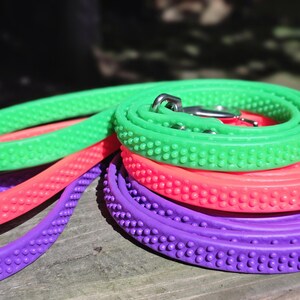 May include: A close-up of several colorful dog leashes. The leashes are in shades of green, pink, and purple. Each leash has a textured surface with small, rounded bumps. A silver metal clasp is visible on the green leash.