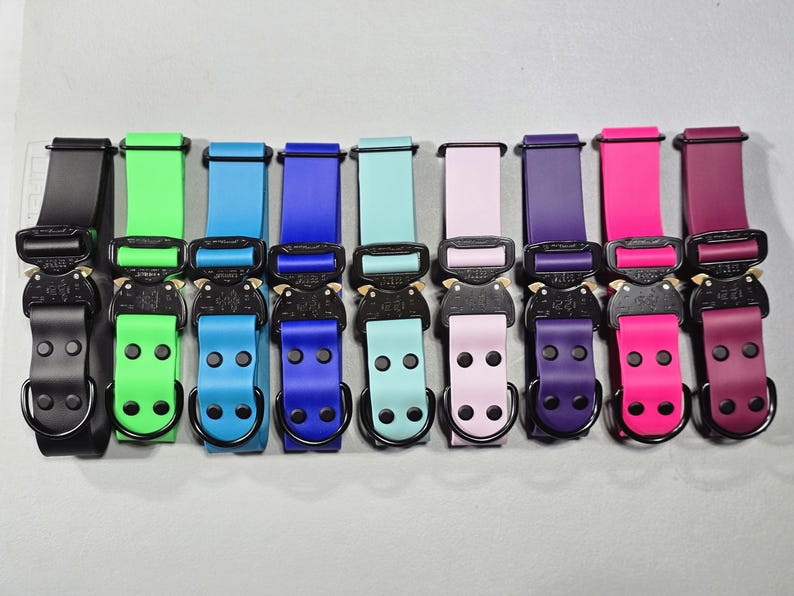 May include: A collection of colorful dog collars with black metal buckles and D-rings. The collars are in various shades, including black, green, blue, pink, purple, and burgundy. The collars are arranged in a row.