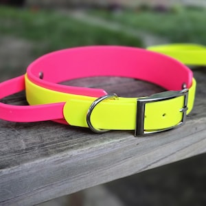 May include: A vibrant pink and yellow dog collar with a silver buckle. The collar is made of a flexible material and has a matching handle. The collar is resting on a weathered wooden surface.