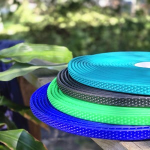 May include: A stack of colorful, textured rolls in shades of blue, green, and black. The rolls have a dotted pattern and are stacked on a wooden surface. The background is blurred, with green foliage.