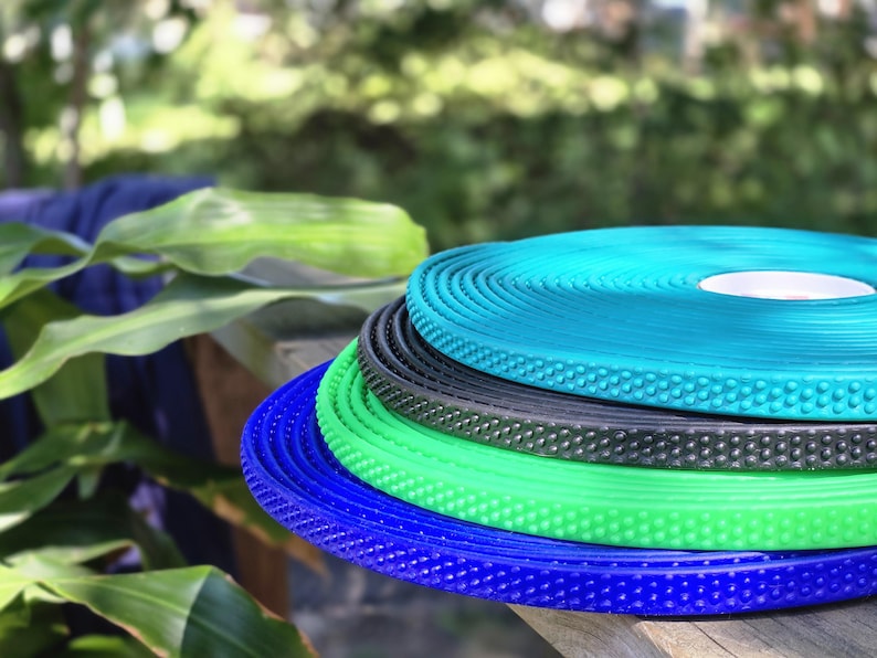 May include: A stack of colorful, textured rolls in shades of blue, green, and black. The rolls have a dotted pattern and are stacked on a wooden surface. The background is blurred, with green foliage.