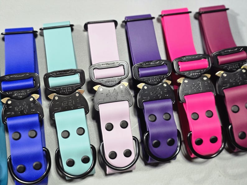 May include: A collection of colorful dog collars in various shades, including blue, teal, pink, purple, and burgundy. Each collar features a black metal buckle and a matching D-ring for leash attachment. The collars are made of a smooth, durable material.