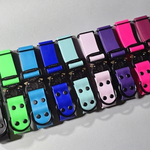 May include: A collection of colorful dog collars in various shades, including black, green, blue, pink, and purple. Each collar features a black metal buckle and matching hardware. The collars are arranged in a neat, organized row.