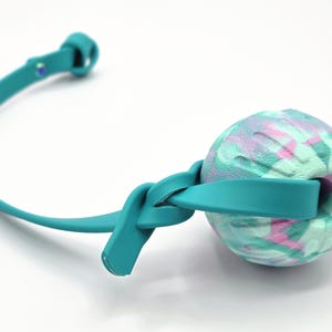 Op de afbeelding: Een turquoise honden speeltje met een getextureerde, veelkleurige bal en een bijpassende riem. De bal heeft een wervelpatroon van roze, groen en wit. De riem is om de bal geknoopt en heeft een lus aan het uiteinde.