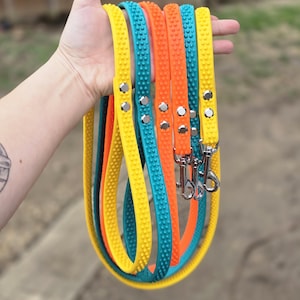 May include: A hand holds several colorful dog leashes. The leashes are yellow, teal, and orange, each with a textured surface and silver hardware. The leashes are made of a flexible material and are designed for pet owners.